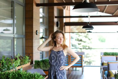 Yükseltilmiş el ile yeşil bitkiler yakınındaki kafede duran genç kadın.