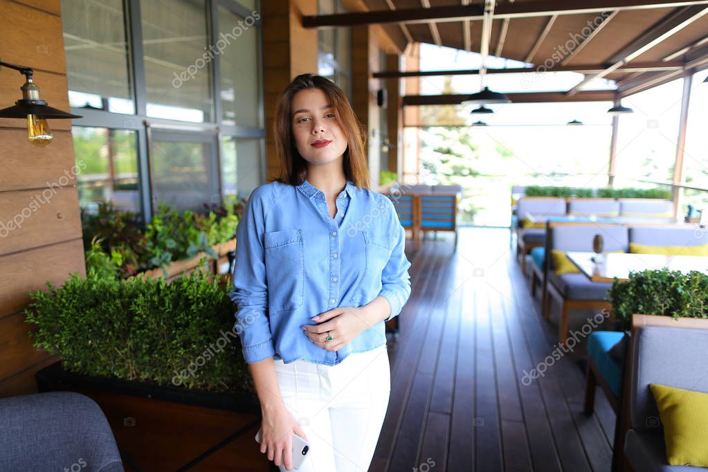 Caucasian woman standing at cafe with smartphone and wearing jeans shirt and white pants.