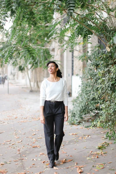 Mujer negra caminando cerca del edificio y árboles verdes, vistiendo blusa blanca y pantalones negros . — Foto de Stock