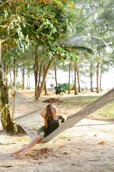 Молодая девушка в платье сидит босиком на плетеном гамаке, песок и деревья на заднем плане . — стоковое фото