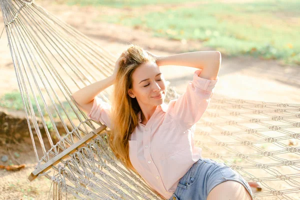 Jovem loira mulher deitada na rede de vime branco na areia, vestindo shorts jeans . — Fotografia de Stock