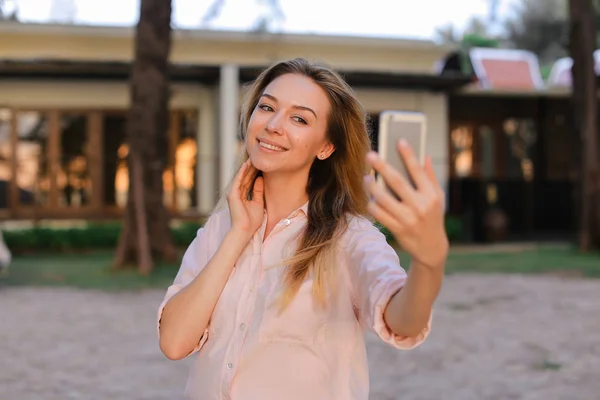 Young pretty woman making selfie by smartphone outside, house in background.