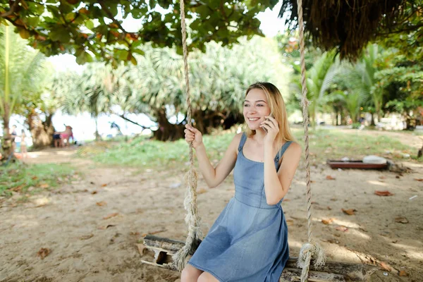 Jovem mulher feliz montando swing e falando por smatrphone, areia e árvore no fundo . — Fotografia de Stock
