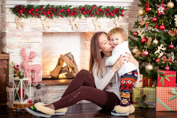 Joven madre sentada con su hija pequeña cerca de la chimenea decorada y el árbol de Navidad . — Foto de Stock