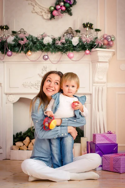 Joven madre caucásica sentada con su hija pequeña cerca de la chimenea de Navidad . — Foto de Stock