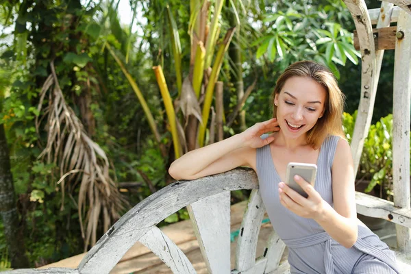 Joven mujer jinete usando teléfono inteligente con palmas en el fondo, sentado en el columpio . — Foto de Stock