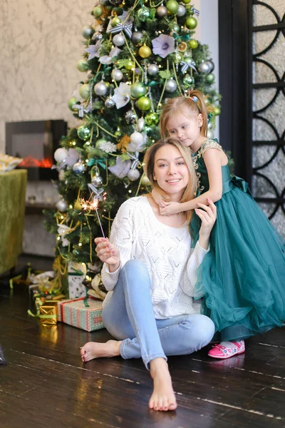 Bonita mujer con luz de bengala usando jeans y sentada con su hija cerca del árbol de Navidad . — Foto de Stock