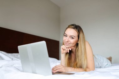 Dizüstü bilgisayar kullanarak ve sabah yatakta yatarken genç mutlu kadın kişi.