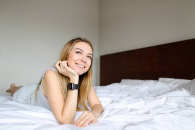 SmartWatch kullanarak ve beyaz keten ile sabah yatakta yatarken genç gülümseyen kadın.