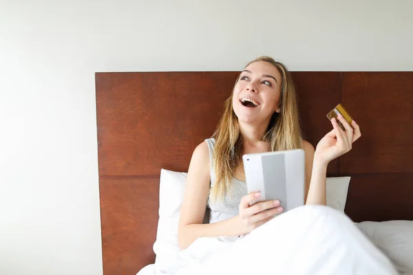 Молода усміхнена жінка йде за покупками онлайн з планшетом і дебетовою карткою, сидячи в ліжку . — стокове фото
