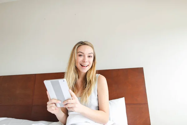 Fiatal mosolygó nő segítségével tabletta, a szociális hálózatok, ült az ágyban. — Stock Fotó