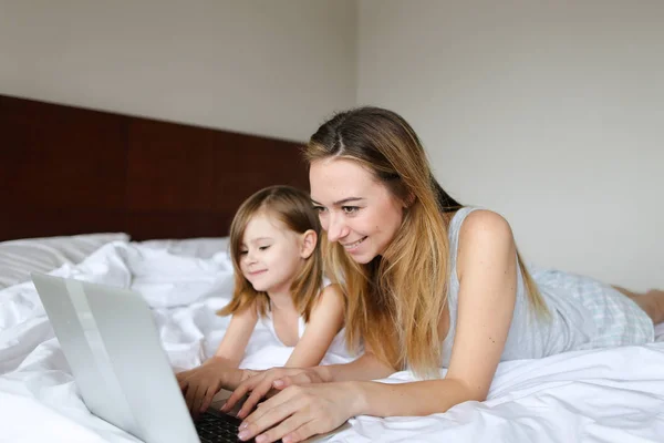 Giovane donna sorridente sdraiata a letto con la piccola figlia al mattino e utilizzando il computer portatile . — Foto Stock