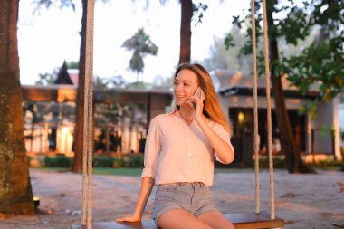 Smartphone ile konuşmak ve oturan genç kadın salıncak, arka planda kum.