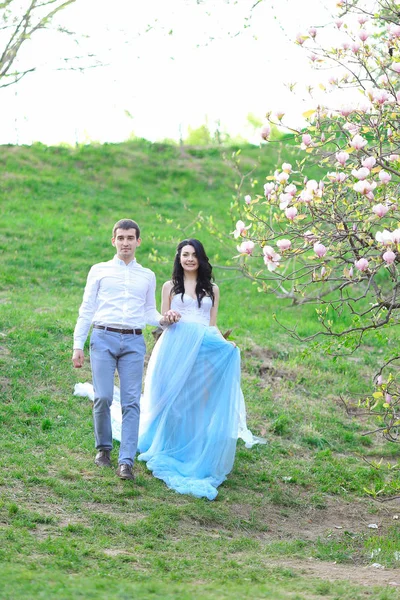 Μελαχρινή νεαρή γυναίκα φοράει μπλε φόρεμα και το περπάτημα κοντά μανόλιας με φίλο. — Φωτογραφία Αρχείου