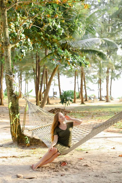 Молодая милая девушка в зеленом платье сидит на плетеном гамаке с песком и пальмами на заднем плане . — стоковое фото