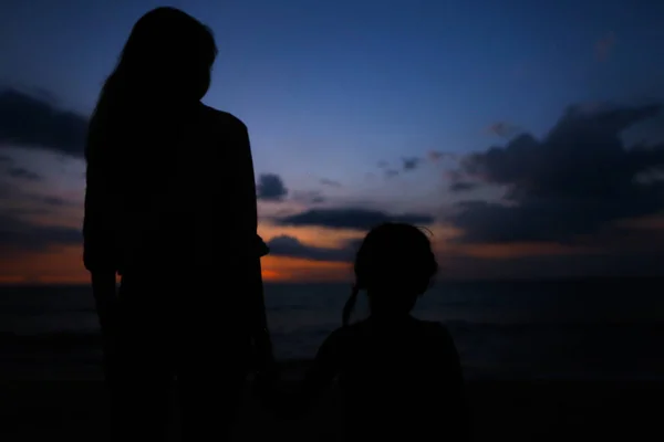 Black silhouette of young woman holding daughter hand and walking on sea shore with sunset backgound. — Stock Photo, Image
