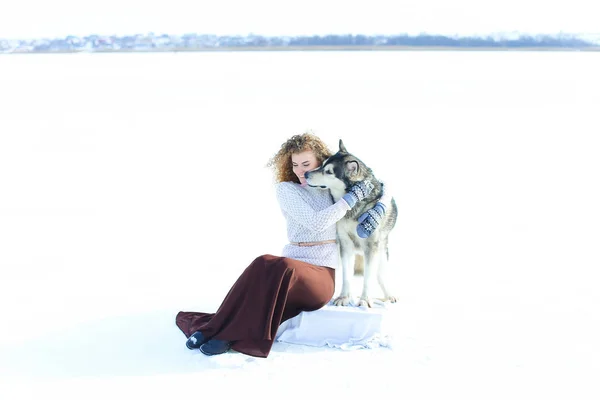 Kobieta nosi sweter, brązowy spódnica i rękawiczki, siedząc na śniegu i przytulanie pies husky. — Zdjęcie stockowe