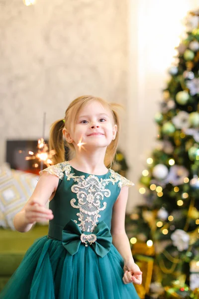 Pequeña chica caucásica vistiendo vestido y de pie con luces de bengala cerca del árbol de Navidad . — Foto de Stock