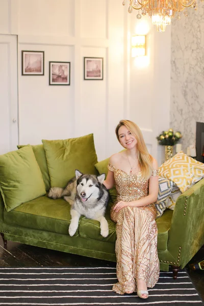 Mooie jongedame dragen jurk zittend op de Bank in de woonkamer met malamute. — Stockfoto