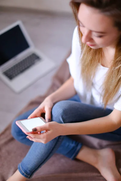Fiatal nő dolgozik a smartphone és ül az ágyon, laptop-ban háttér. — Stock Fotó