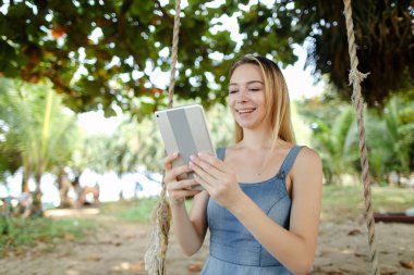 Kot sundress giyen tablet kullanma ve salıncak kum üzerinde sürme genç mutlu kadın.