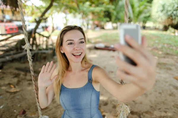Genç kız sürme salıncak gülümseyen ve selfie smartphone, kum ve ağaçlar tarafından arka planda yapma. Yaz tatillerini ve modern technlogy, boş zaman kavramını.