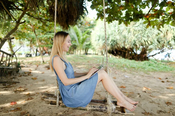 Jovem caucasiana trabalhando com laptop e montando no balanço, areia no fundo . — Fotografia de Stock