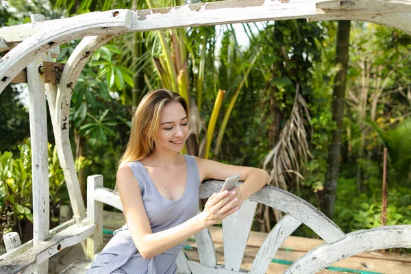 Salıncakta sürme ve egzotik Bahçe, avuç içi arka plan smartphone kullanarak genç kafkas kadın.