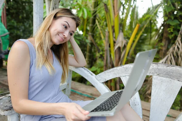 Happy woman using laptop and sitting in exotic garedn with palms in background. — Stock Photo, Image