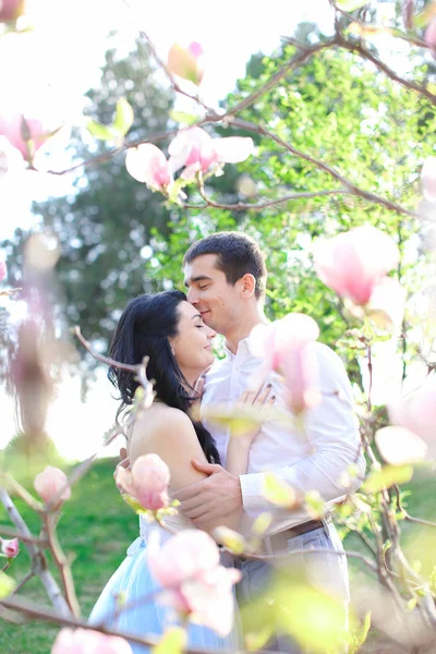 Morena menino abraçando e beijando menina caucasiana perto de magnólia . — Fotografia de Stock