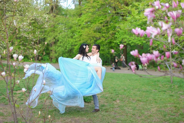 Homem segurando mulher vestindo vestido azul no parque perto de magnólia árvore . — Fotografia de Stock