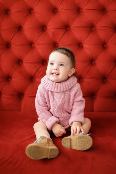 Liten söt kvinnlig baby sitter i röd bakgrund på soffa. — Stockfoto