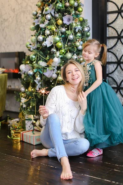 Joven mujer bonita con luz de bengala usando jeans y sentada con su hija cerca del árbol de Navidad . — Foto de Stock