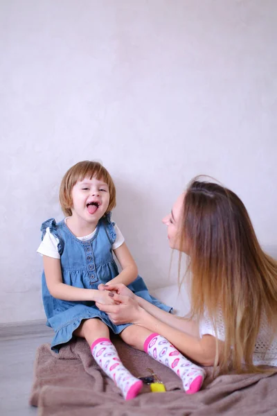 Young blonde mother playing with little daughter and sitting on bed. — Stock Photo, Image