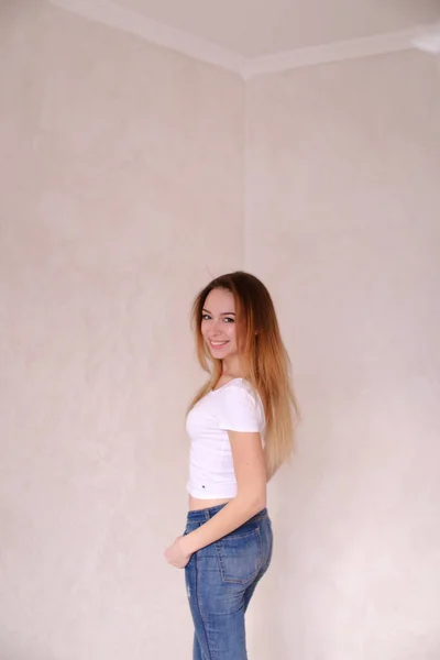 Young blonde woman wearing white shirt and jeans standing near wall. — Stock Photo, Image
