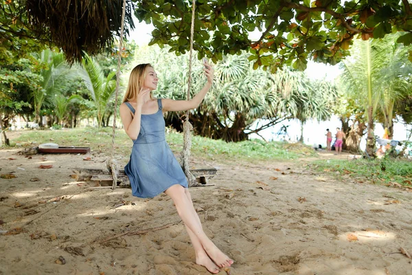 Salıncak sürme ve selfie smartphone, kum ve ağaçlar tarafından arka planda yapma genç beyaz kız.