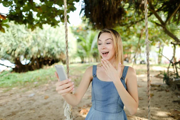Salıncak kum üzerinde smartphone kullanmasına ve sosyal ağlar, Genç neşeli kız sürme.