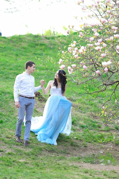 Jonge brunette meisje draagt blauwe jurk en wandelen in de buurt van magnolia met vriendje. — Stockfoto