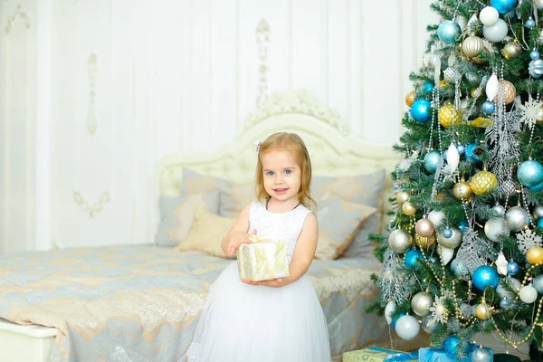 Kleines Mädchen in weißem Kleid hält Geschenk in der Nähe geschmückten Weihnachtsbaum im Schlafzimmer. — Stockfoto