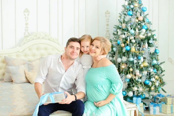 Kleine kaukasische Tochter sitzt mit glücklichem Vater und schwangerer Mutter auf Bett neben geschmücktem Weihnachtsbaum. — Stockfoto