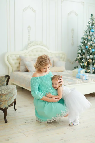 Schwangere junge Frau trägt blaues Kleid, umarmt Bauch und sitzt mit kleiner Tochter neben Weihnachtsbaum im Schlafzimmer. — Stockfoto