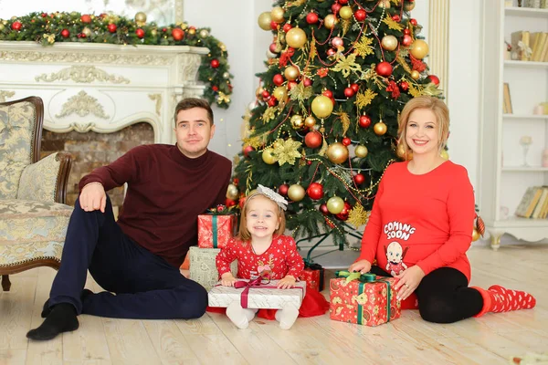 Kleine Tochter sitzt mit Vater und schwangerer Mutter am Weihnachtsbaum und bewahrt Geschenke auf. — Stockfoto