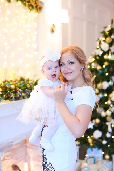 Jovem mãe vestindo vestido branco e mantendo pouco bebê feminino perto da árvore de Natal . — Fotografia de Stock