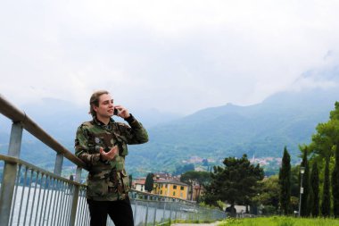 Avrupa delikanlı yanında tırabzan, Como Gölü ve Alpler Dağı arka planda smartphone konuşuyor.