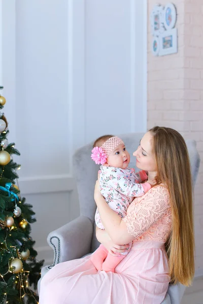 Noel ağacı yakınındaki pembe elbise giyen çocuk ile kol sandalyede oturan genç güzel kadın.