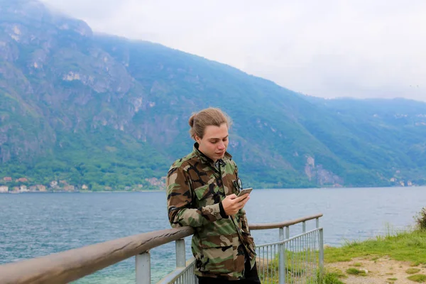 Joven con teléfono inteligente cerca de barandilla, lago Como y la montaña de los Alpes en el fondo . — Foto de Stock