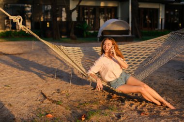 Hamakta oturmuş ve smartphone, yaz tatillerini konuşan genç kadın.