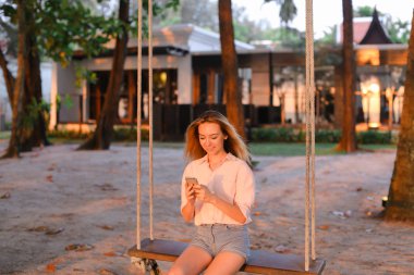 Genç smartphone kullanan kadın güzel ve salıncakta, oturan arka planda kum.