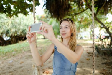Genç kız salıncak sürme ve selfie smartphone, kum ve ağaçlar tarafından arka planda yapma sevindirmek.