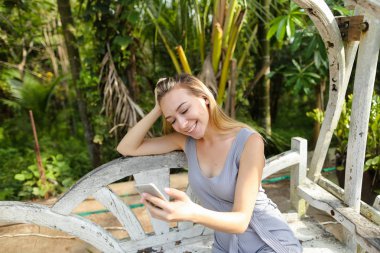 Avuç içinde geçmiş, salıncakta oturan Smartphone kullanan genç mutlu kız. Modern teknoloji ve egzotik tatil dinlenme kavramı.